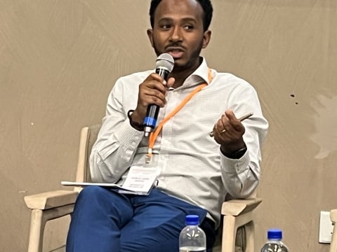 A dark-skinned man with black hair and a beard, wearing a white shirt and jeans and an orange lanyard around his neck, speaks into a microphone while sitting in a chair. 