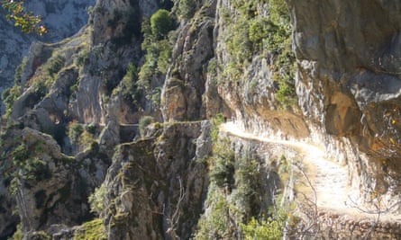 Hikers walk along a steep mountainside path, illuminated in bright sunshine.