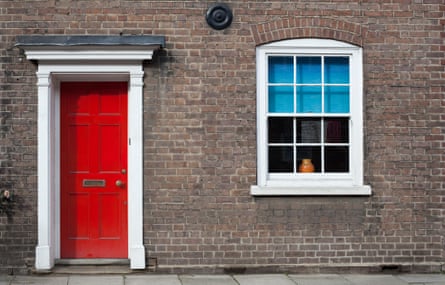 A red door, a window with blue blinds, and white surrounds on a brick townhouse in a British village.