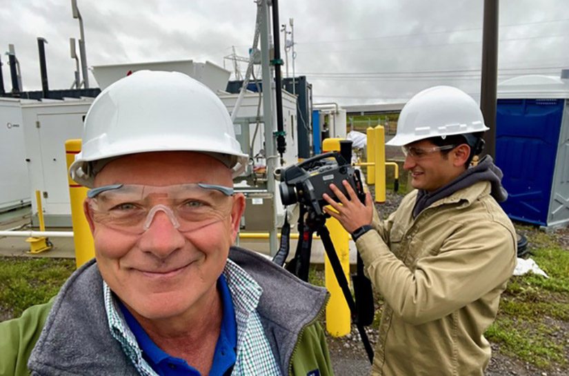 A man faces the camera with another man looking left in the background using a camera on a tripod.