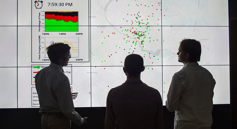 Three men with their back turned away from the camera look at a large screen.