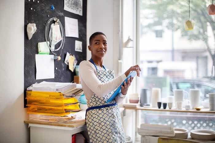 A woman wearing an apron and standing in her small pottery shop holding a clipboard.