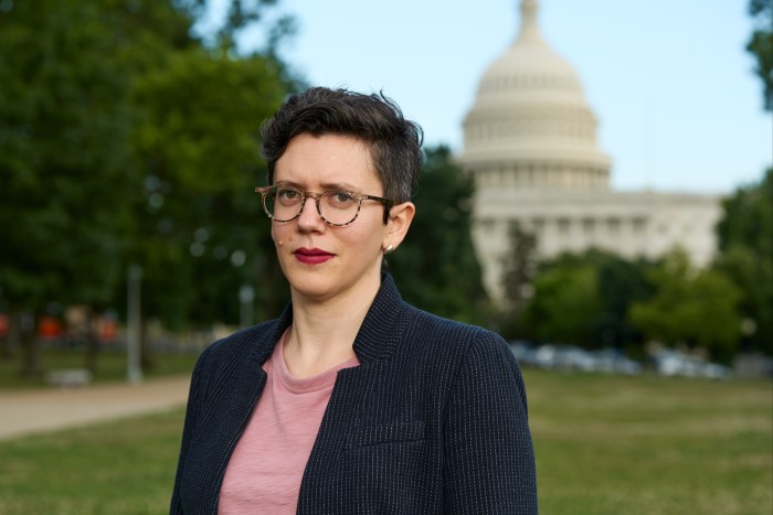 A short-haired woman with glasses and in office wear poses in front of a classically designed building with a dome