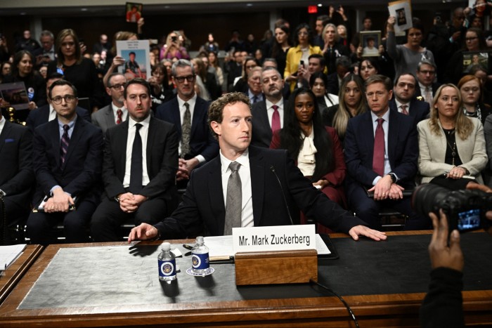 Mark Zuckerberg wearing a suit and tie, sitting at a desk with rows of people in smart clothes apparently listening