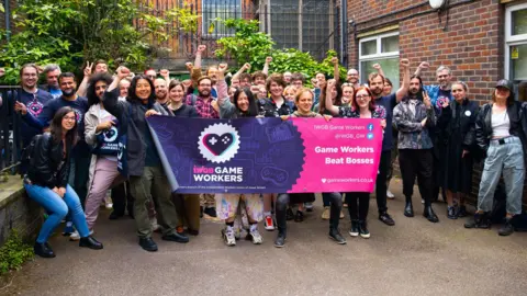 IWGB A group of about 30 casually dressed people standing in the courtyard of an office building. Four people at the front are holding a purple and pink banner with the IWGB union Game Workers' branch logo and the slogan "Game Workers Beat Bosses". Some members of the group have their clenched fists raised above their heads.