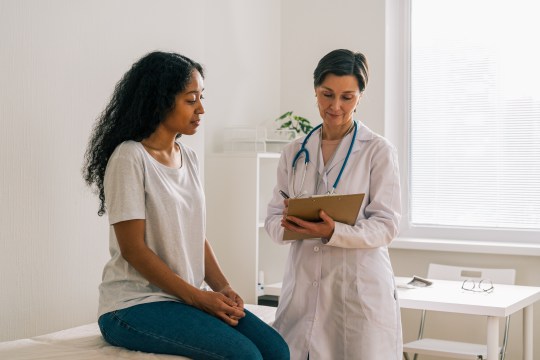 A patient sat with a doctor holding a clipboard in a GP's office.