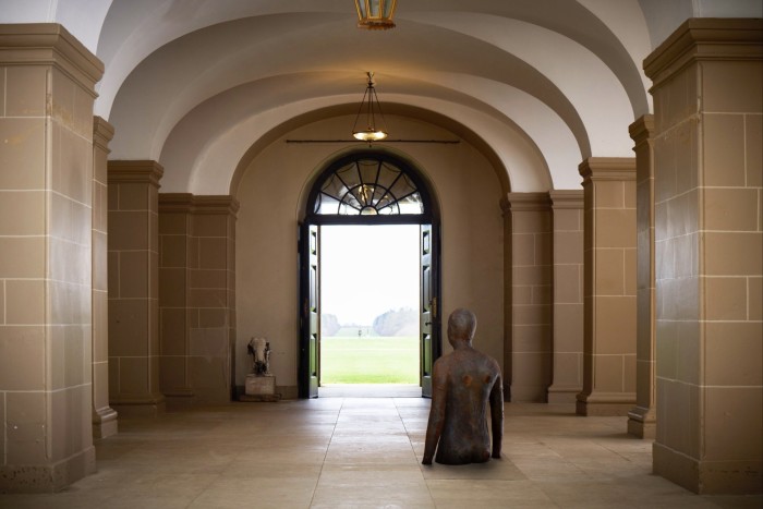 A statue of a person up to their waist in a hall way with arches that looks out through a door frame to a grassy field with another statue in the distance