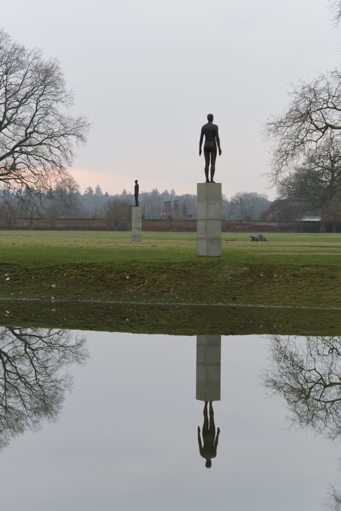 A large cast-iron statue of a person on a plinth reflected in a pool of water. There are trees either side