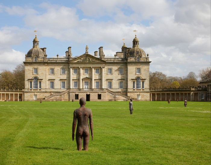 Cast-iron statues of people in a grassy field with a manor house in the background. The statue in the foreground looks has his feet buried in the ground