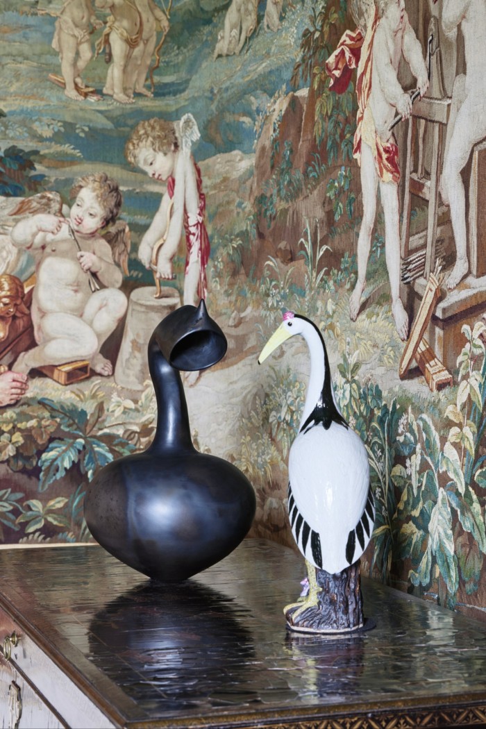 Two sculptures placed on a table in front of an old tapestry. One sculpture is a detailed ceramic figure of a white bird, the other is a dark vase with a long neck