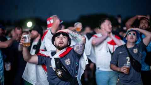 Young men in blue or white football shirts, some of the draped in England flags, look up tensely. Some of them hold a hand to their head