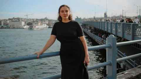 Natalia Sindeyeva has long dark hair and a dark dress. She leans on a railing with a river behind her