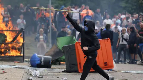 Reuters A man in a black hoodie with his face covered by sunglasses and a balaclava throws a stick, while in the background are overturned bins, a fire and a large crowd of people