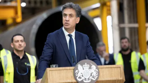 Reuters Energy secretary Ed Miliband stands behind a lectern with a government crest and speaks to workers at an industrial site 