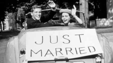 1950s shot of a couple with a ‘Just Married’ banner on the back of their car