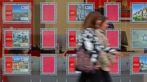 Pedestrians walk past the front window of an estate agent