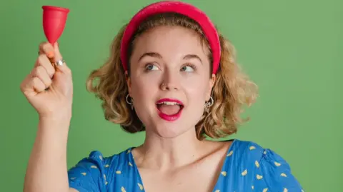 Instagram/Bella Humphries Comedian Bella Humphries wearing a red headband with mouth slightly open  holding a mooncup to the left and looking up towards it. 