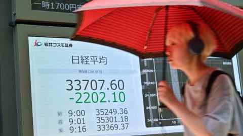 A pedestrian holding an umbrella walks past display boards showing numbers on the Tokyo Stock Exchange