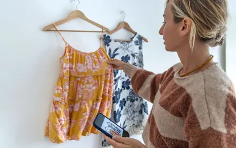 Getty Images A woman adjusts a dress hanging on a wall in her home, while she holds her phone with the camera app open