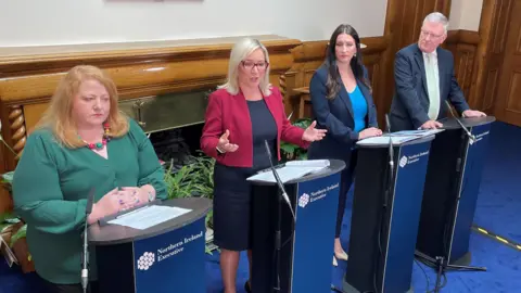 PA Media Naomi Long, Michelle O'Neill, Emma Little-Pengelly, and Mike Nesbitt standing at podiums with microphones. Michelle O'Neill is speaking. They are standing in a room with wood panelling in the background.