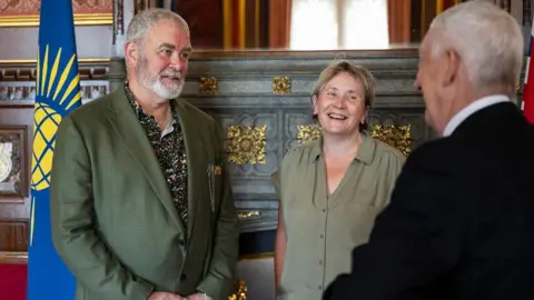 Terry Wiggins, wearing a green blazer and patterned shirt, faces the camera, as Lindsey Hoyle (seen from behind) talks to him 