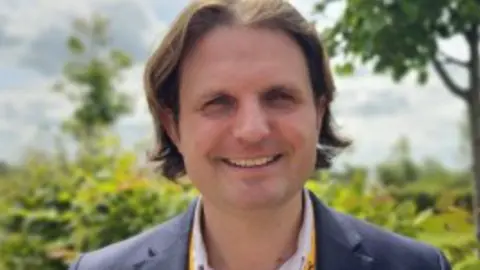 Steff Aquarone, MP for North Norfolk, has mid-length brown hair parted down the middle. He's smiling on a sunny day, wearing a navy suit and a pale shirt with the neck open.