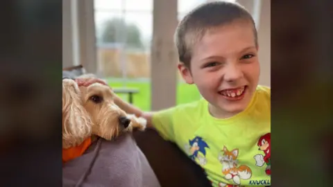 Julie Tipping A young boy with blonde hair is laughing and looking into the camera. He is wearing a neon t-shirt with cartoon characters printed on the front. Beside him is a light brown dog lying on the back of a grey sofa. 