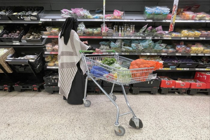 A shopper pushes a shopping cart filled with groceries in front of the produce section in an Asda supermarket