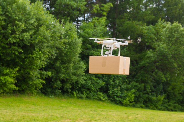 A small white drone carrying a brown cardboard boxer bigger than itself over grass next to full-leaved bushes and trees.