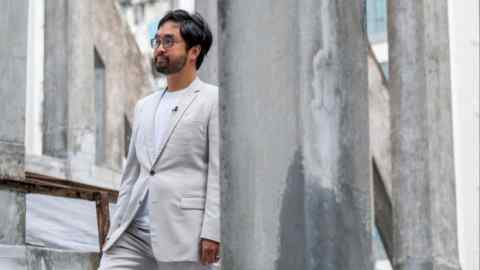 Adrian Cheng standing outdoors in front of a concrete structure at the State Theatre in Hong Kong. He is wearing a light-colored suit and glasses.