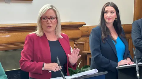 PA Media Michelle O'Neill and Emma Little-Pengelly standing behind podiums with microphones. Michelle O'Neill is speaking. They are standing in a room with wood panelling in the background