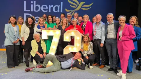 BBC Liberal Democrat MPs pose happily around a giant 72 sign, made of lightbulbs 