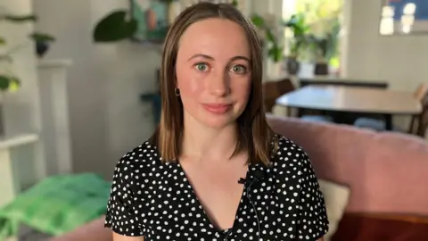 Liz is a woman with shoulder-length brown hair. She is sat in a living room with plants and furniture in the background. She is wearing a black and white spotty outfit.