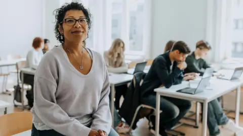 Getty Images Teacher sits on a desk in a classroom with pupils working at desks behind her.