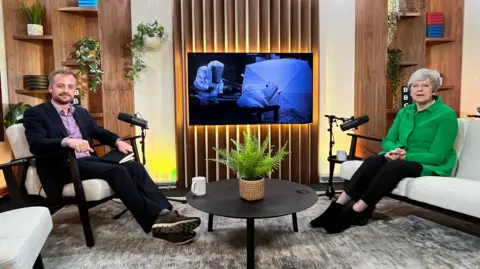 Former Prime Minister Theresa May sitting on a sofa opposite the BBC's Dan O'Brien. In between them is a low coffee table with a plant on it. There are mics set up next to both of them and a picture is shown on a TV on the wall of men in white suits taken during the chemical attack in 2018.