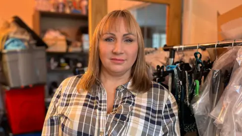 Lorna Edgar Woman with shoulder length blonde-hair in checked shirt smiling at the camera. She is next to a clothes rail with sparkly outfits.