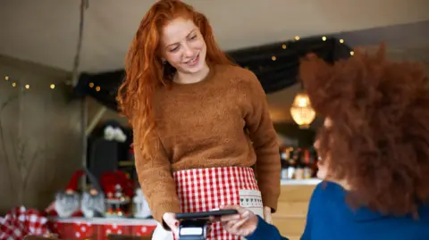 Getty Images Customer in cafe making contactless payment with mobile phone to a waitress wearing a red and white checked apron