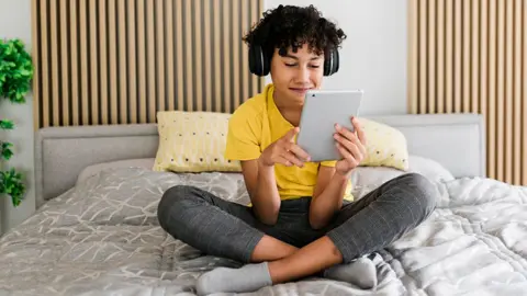 Getty Images A young person wearing jeans and a yellow t shirt uses a tablet with headphones on while sitting cross-legged on a bed with grey sheets on