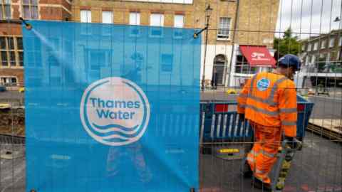 A sign displaying the Thames Water logo is attached to protective barriers at a water supply worksite in London. A worker in an orange high-visibility uniform is seen in the background within the fenced area.