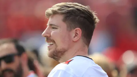 Getty Images MrBeast AKA Jimmy Donaldson pictured at an American Football game. He wears a white football shirt with red numbering on his shoulder. He has a short beard and short sandy hair. 