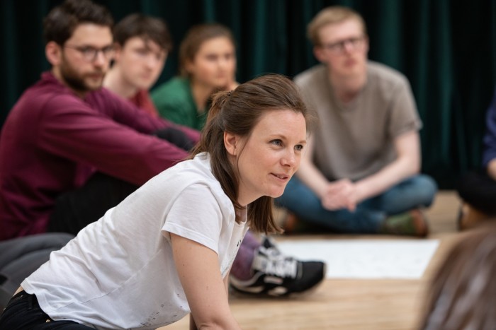 A group of people sit on the floor. In the foreground is a woman with long brown hair, wearing a white T-shirt. She is smiling and has a very focused expression