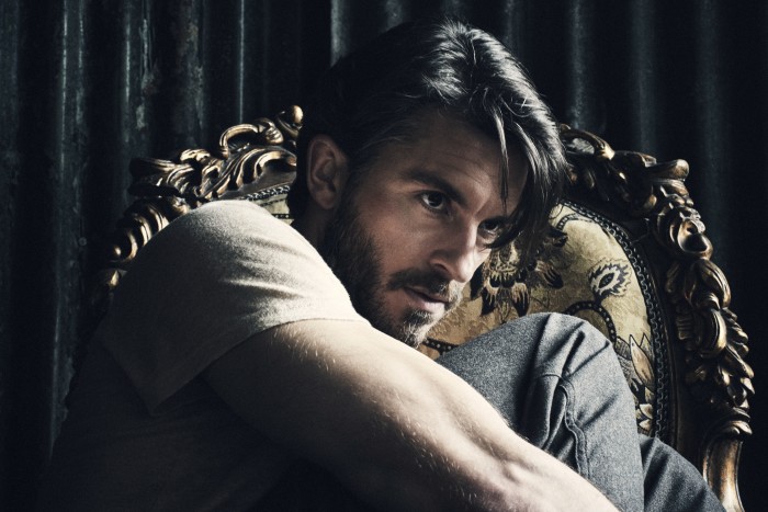 A man with short dark hair and beard sits on an ornate chair with a sultry expression 