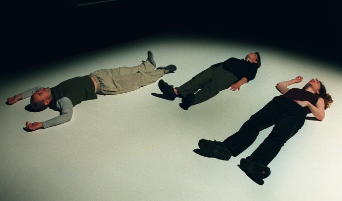 Three people, dressed casually,  lie prone on a stage. Their eyes are open, and staring upwards