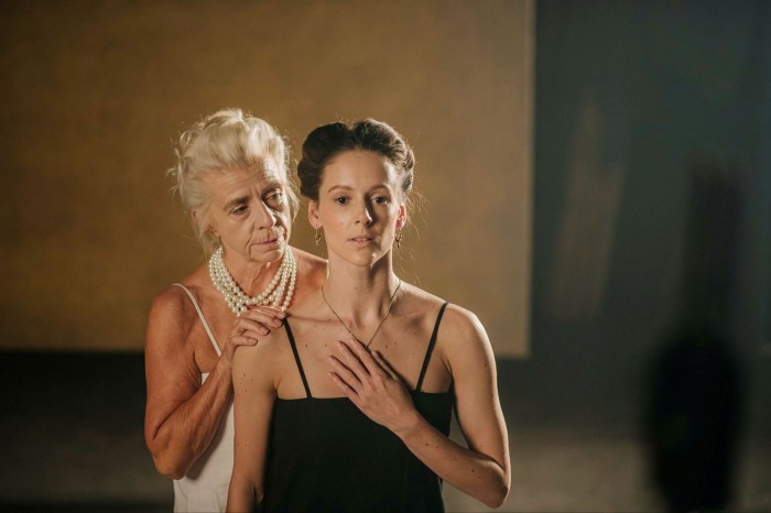 Two women stand on a stage. The older of the two, who has grey hair and is wearing pearls, stands behind the younger woman, with her hands on her shoulder