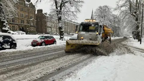 BBC/Yunus Mulla A gritting lorry clears a road in Harrogate with cars parked next to mounted snow on the pavements and hills.