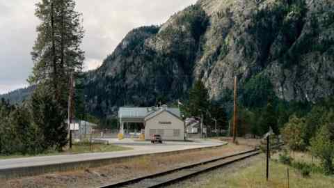 A white building with Cascade written on the front. There is a red truck in front of the building. a railway line in the foreground and rocky mountain outcrop in the background