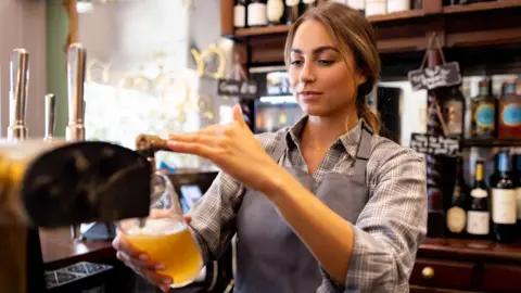 Getty Images Young woman pouring a pint in a pub, wearing a grey checked shirt and plain grey item