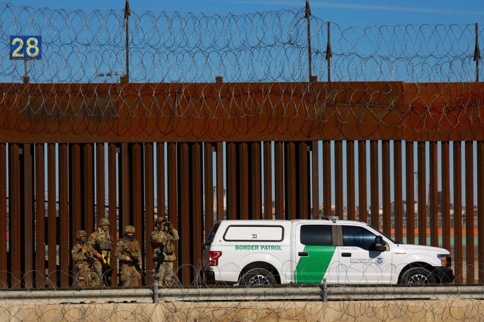 Border Patrol agents standing by a border wall with a Border Patrol vehicle.