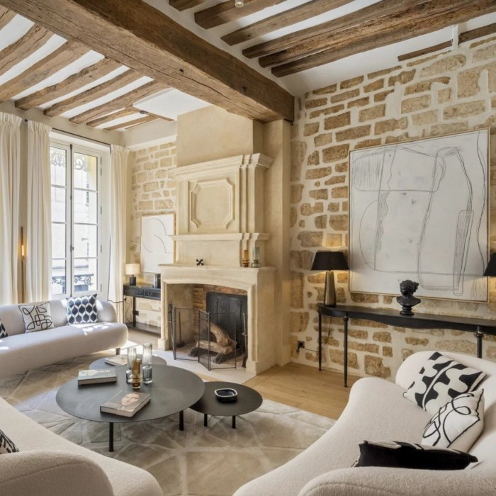 A living room with exposed stone walls, wooden ceiling beams, a grand fireplace, modern white sofas, abstract artwork, and soft natural light streaming through tall French windows