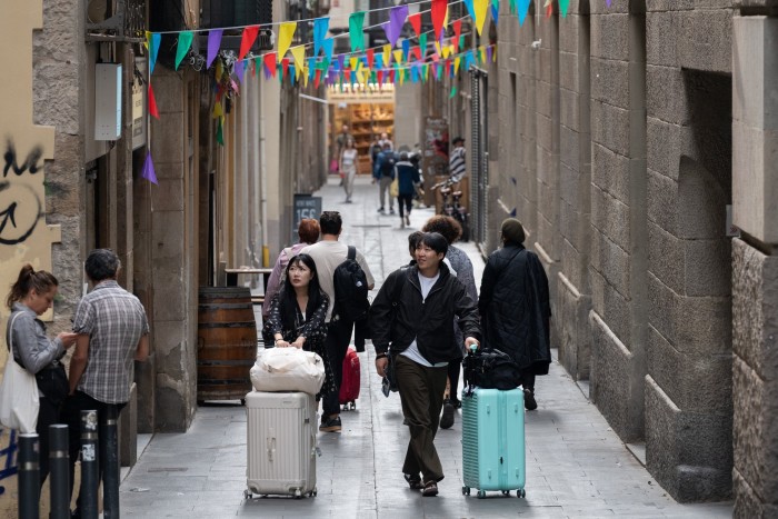 A narrow alley with stone walls and colourful flags overhead, bustling with tourists pulling suitcases and locals walking